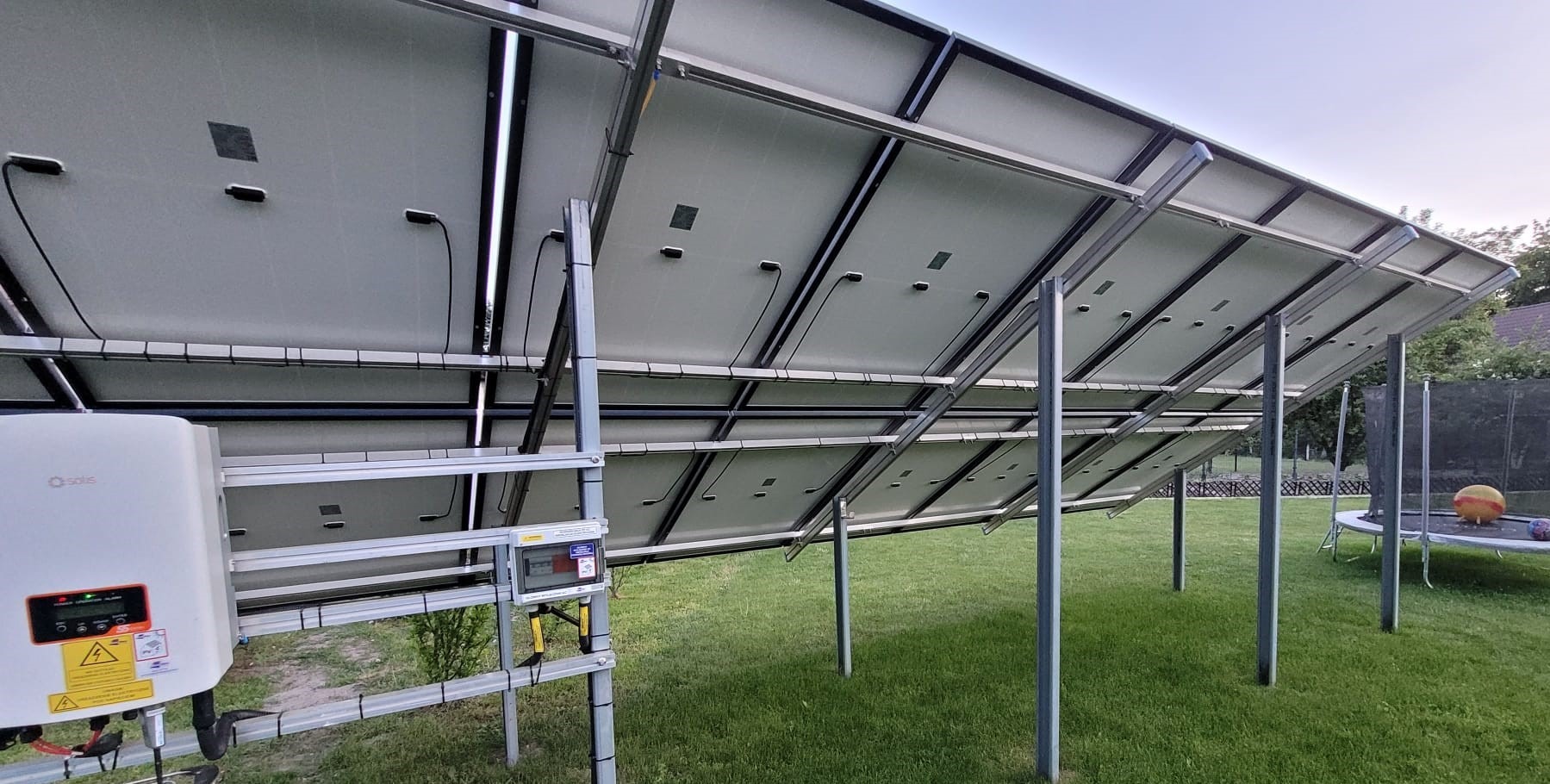 Widok od spodu na panele fotowoltaiczne zamontowane na metalowej konstrukcji na trawniku, widoczny inwerter Solis i skrzynka elektryczna.