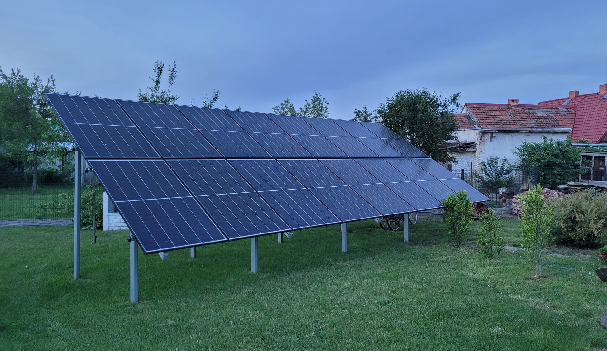 Panel fotowoltaiczny na metalowej konstrukcji, zainstalowany na trawniku w ogrodzie, widoczny rower oparty o konstrukcję, w tle dom z czerwonym dachem.
