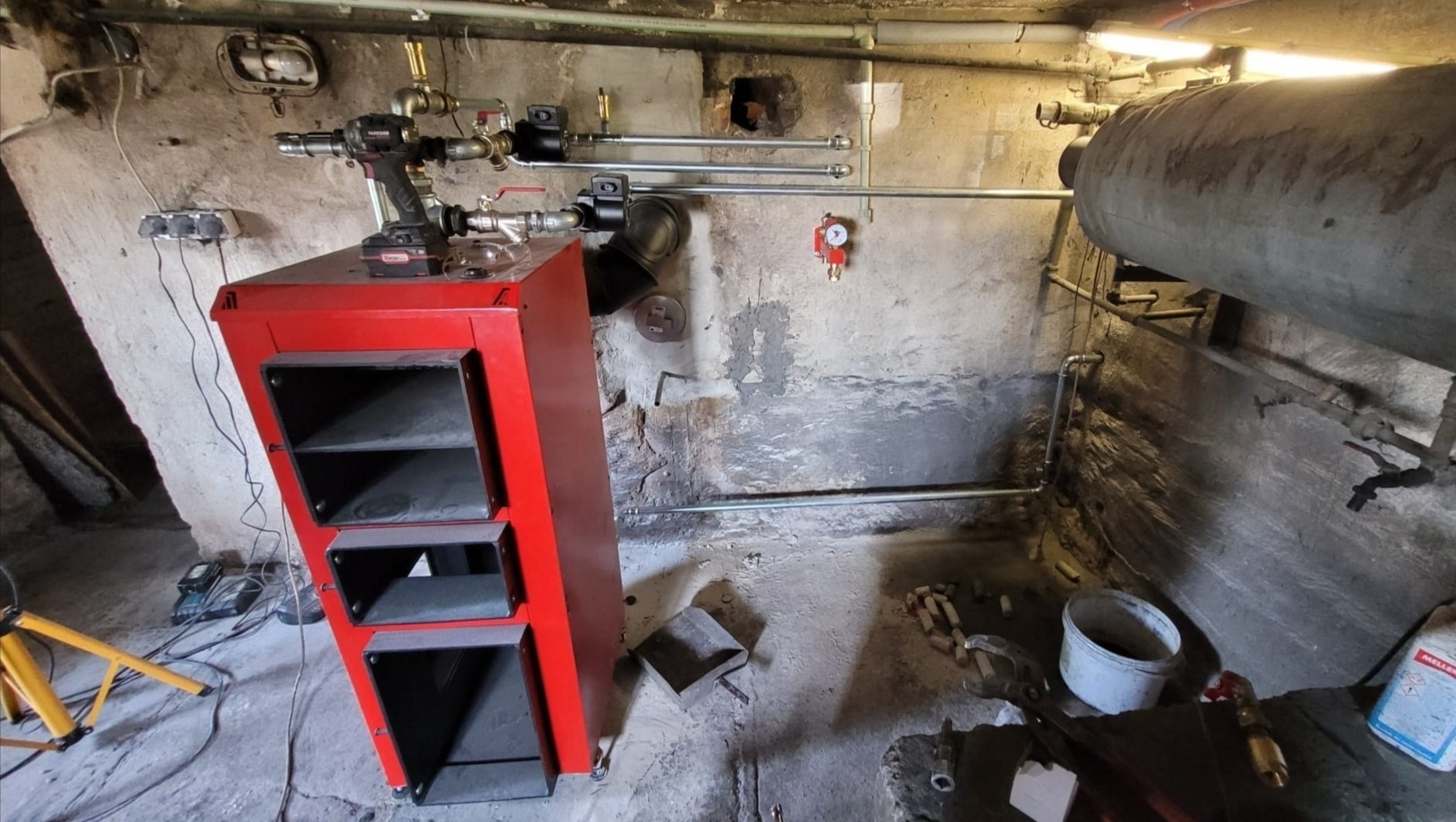 Czerwony piec na paliwo stałe z otwartymi drzwiczkami, zamontowany w piwnicy z widocznymi rurami, narzędziami i elementami instalacji grzewczej.