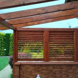 Drewniana pergola z żaluzjami i zadaszeniem, widok na ogród z zielenią, brązowe elementy konstrukcyjne, ceglana podstawa.