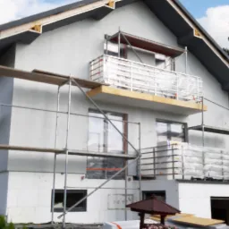 Dom jednorodzinny w trakcie montażu izolacji termicznej ścian zewnętrznych, widoczne rusztowania i zabezpieczona folią balustrada balkonu.