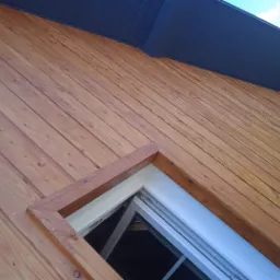 Drewniana elewacja budynku z oknem, widok pod kątem, ciemny dach i fragment nieba w tle.