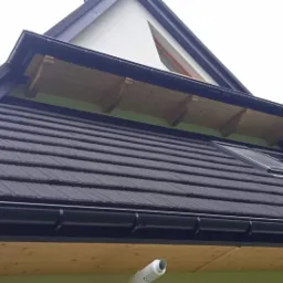 Fragment dachu z czarną dachówką, oknami dachowymi i czarnymi rynnami, widoczna drewniana podbitka i fragment ściany szczytowej.