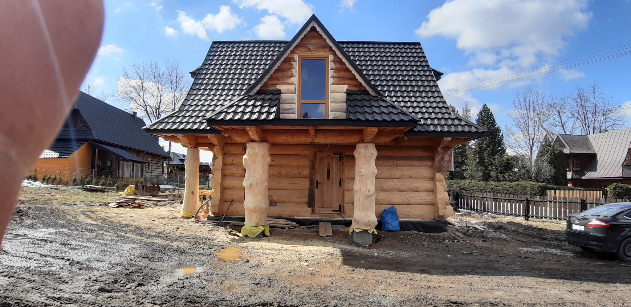 Nowo wybudowany drewniany dom w stylu góralskim z ciemną dachówką, widoczne surowe drewniane kolumny i teren budowy.