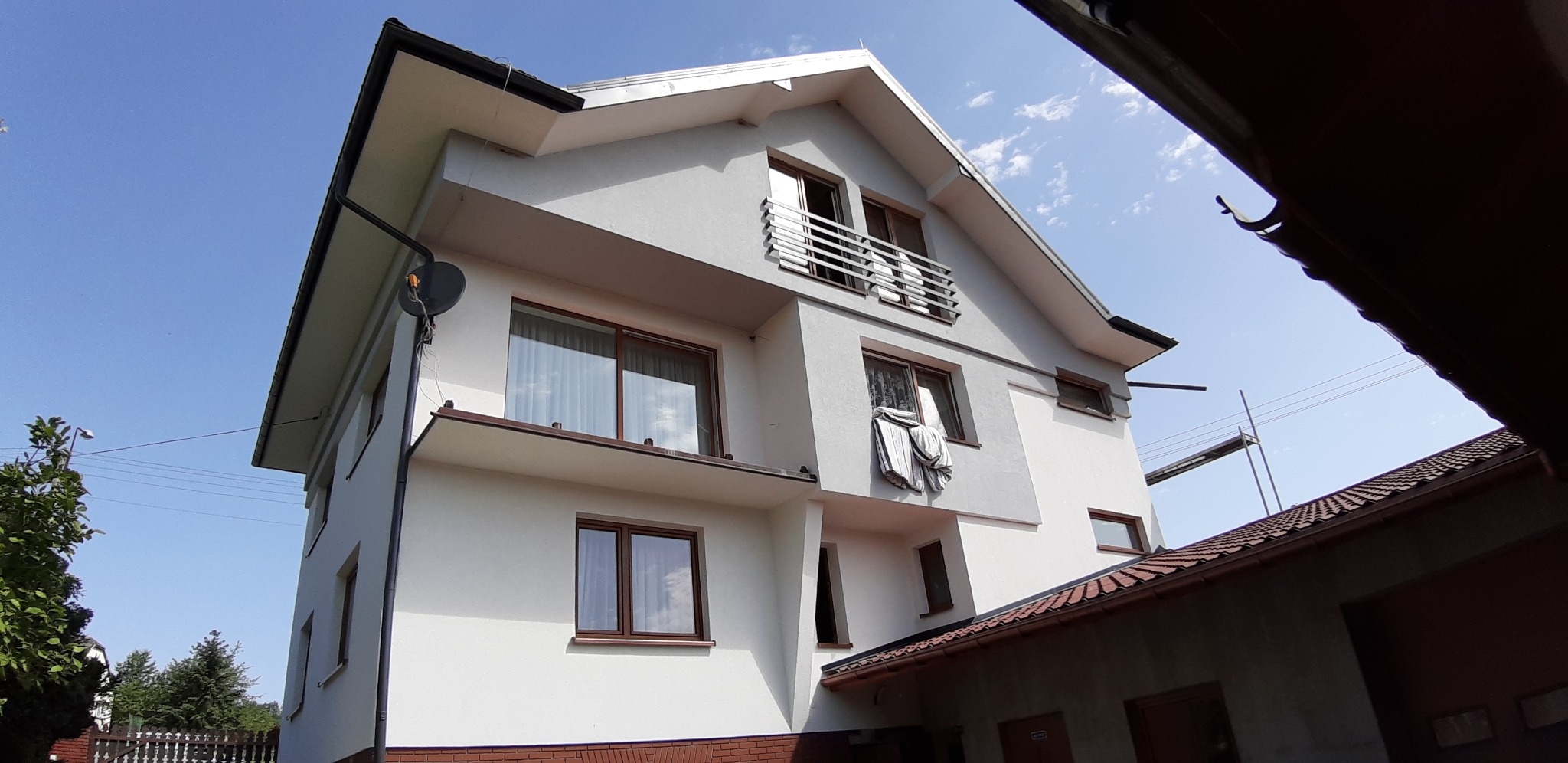 Dwupiętrowy dom z szarą elewacją, brązowymi oknami i czerwono-brązową ceglaną podstawą, widziany z dołu pod kątem, z widoczną anteną satelitarną i suszącym się praniem na oknie.