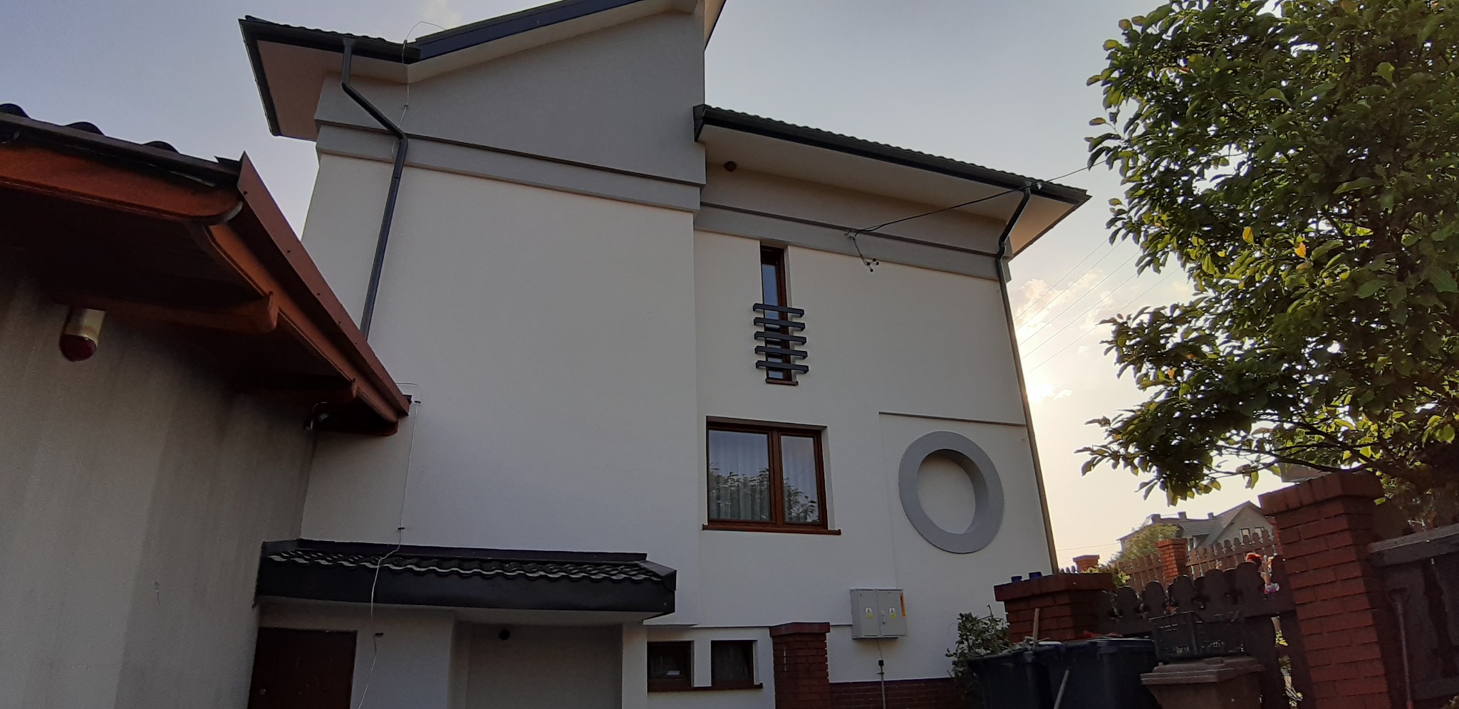 Dwukondygnacyjny dom z jasną elewacją, ozdobiony szarym okręgiem i poziomymi listwami przy oknie, widoczny z perspektywy ogrodu z drzewem.