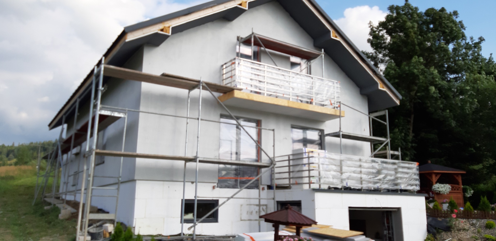 Dom jednorodzinny w trakcie montażu izolacji termicznej ścian zewnętrznych, widoczne rusztowania i zabezpieczona folią balustrada balkonu.