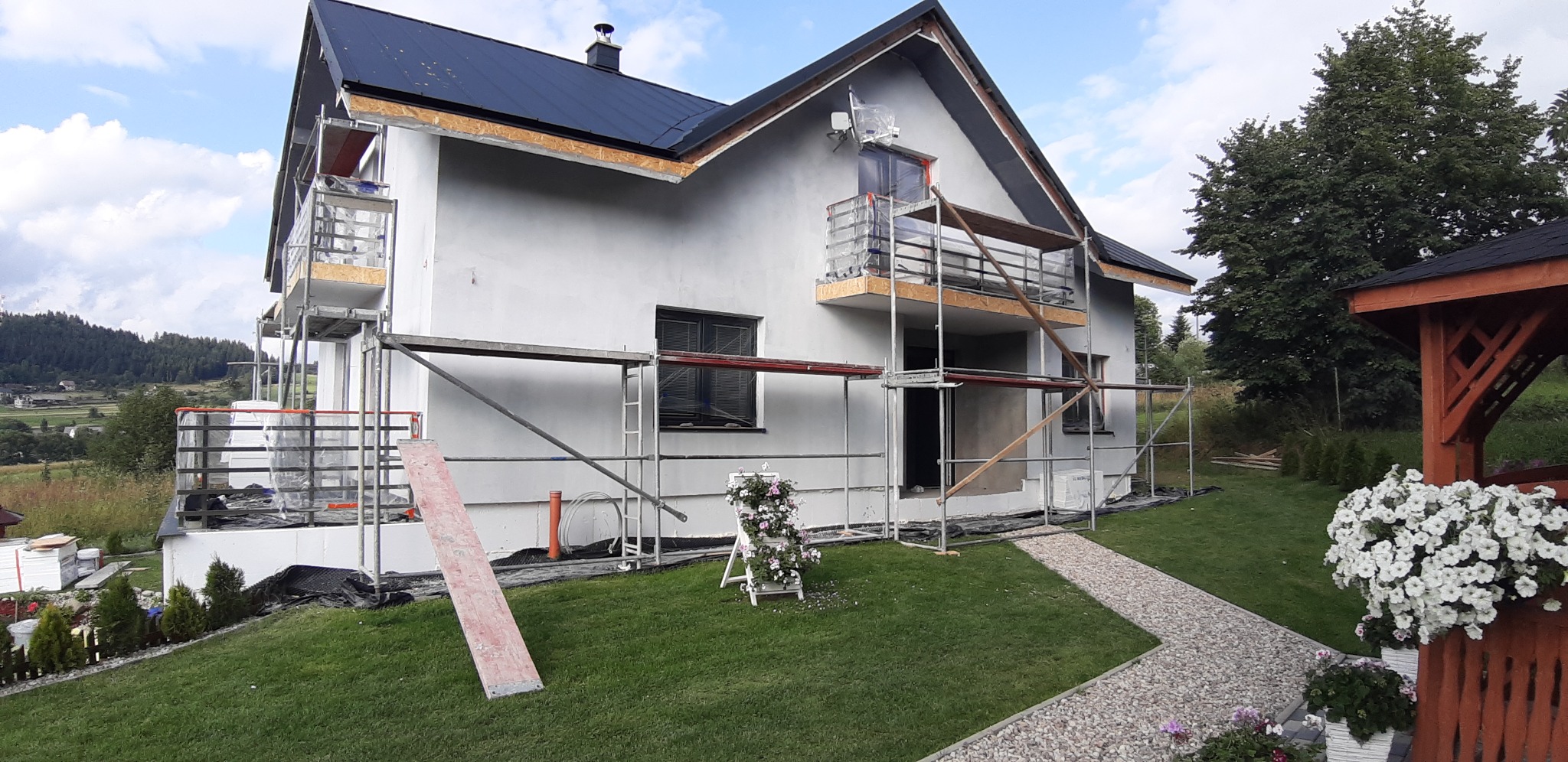 Dom jednorodzinny w trakcie prac elewacyjnych, z widocznymi rusztowaniami i świeżo położonym szarym tynkiem, na tle zielonego trawnika i górskiego krajobrazu.