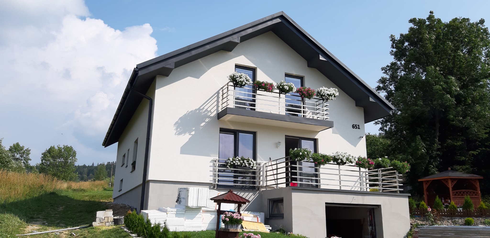 Dwukondygnacyjny dom z białymi ścianami i szarym dachem, balkony z kwiatami, w tle zieleń, na trawniku leżą płyty styropianowe.