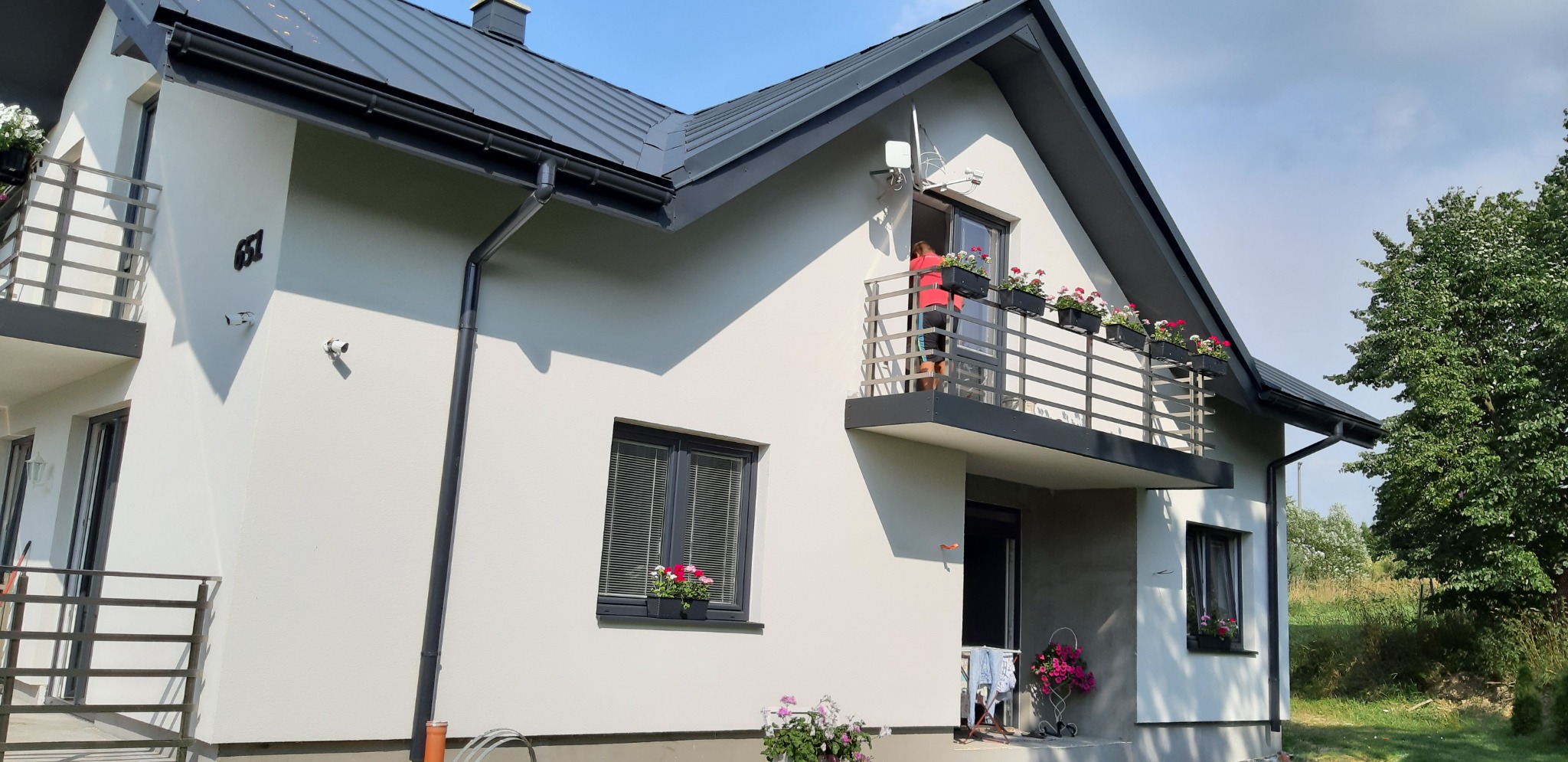 Jasny, otynkowany dom jednorodzinny z ciemną dachówką i oknami, balkon z kwiatami, kobieta w czerwonej bluzce podlewa kwiaty na balkonie.