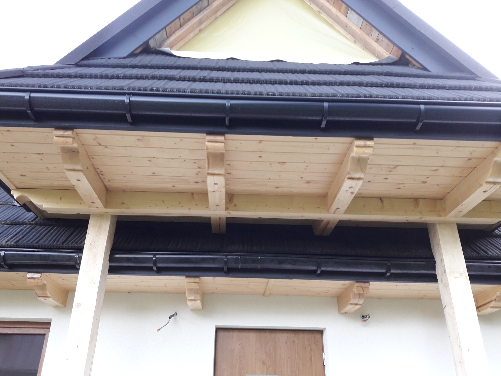 Drewniany podbitka dachowa z elementami zdobniczymi, czarne rynny, fragment elewacji z widocznymi oknami i drzwiami wejściowymi.