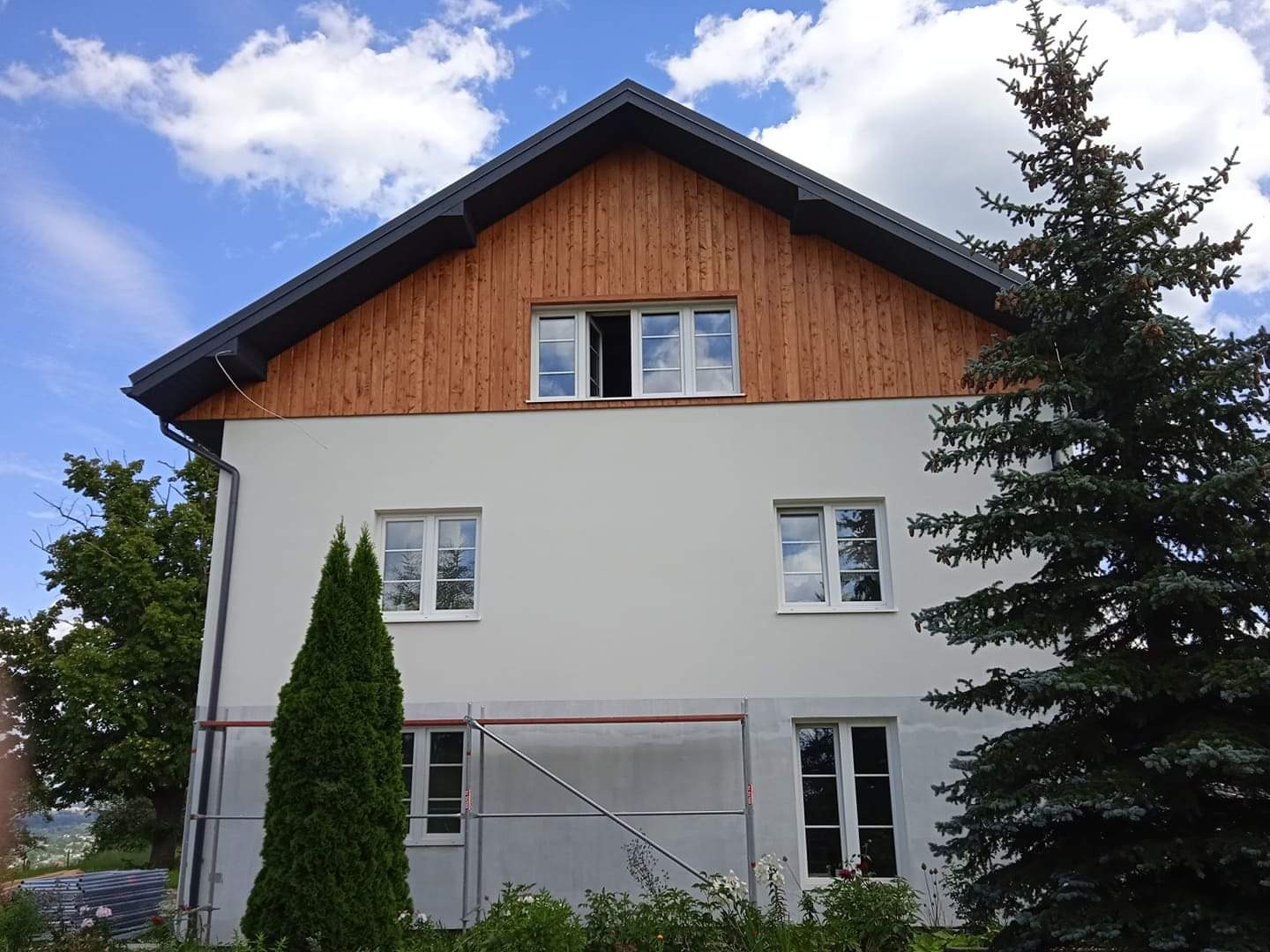 Dwukondygnacyjny dom z jasną elewacją i drewnianą górną częścią, częściowo zakryty rusztowaniem i zielenią, pod błękitnym niebem z chmurami.