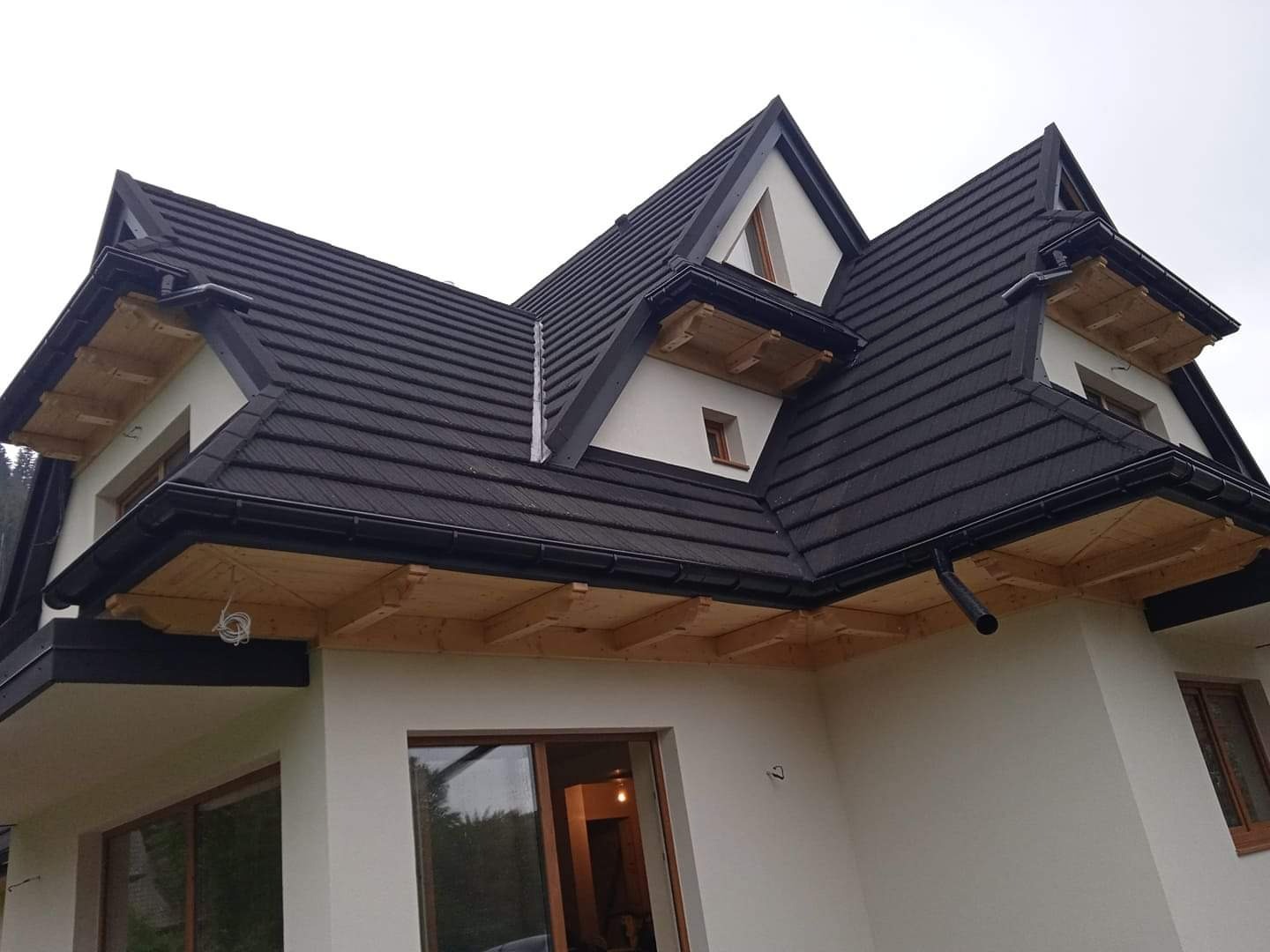 Nowoczesny dom z ciemną, metalową dachówką karpiówką, drewnianymi elementami okapu i białymi ścianami elewacji, widok z dołu.