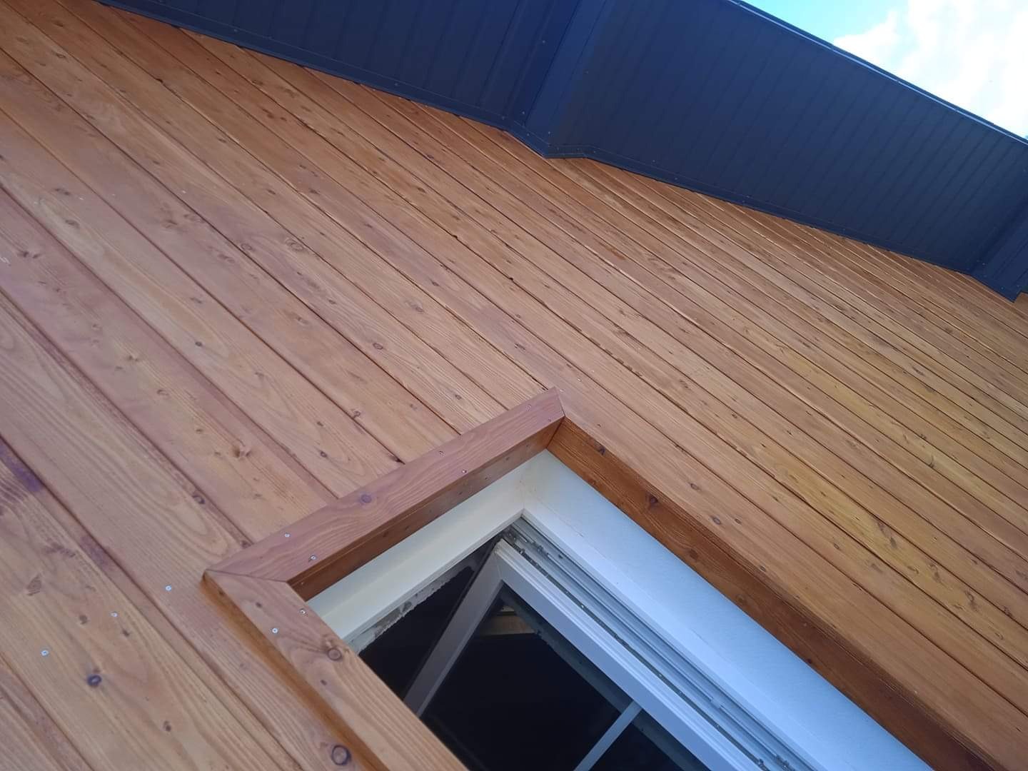 Drewniana elewacja budynku z oknem, widok pod kątem, ciemny dach i fragment nieba w tle.