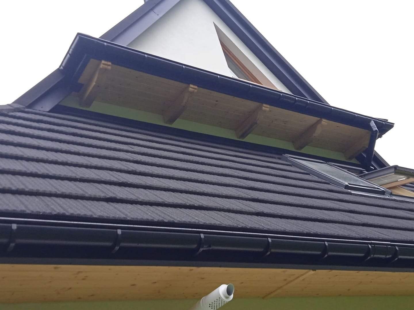 Fragment dachu z czarną dachówką, oknami dachowymi i czarnymi rynnami, widoczna drewniana podbitka i fragment ściany szczytowej.
