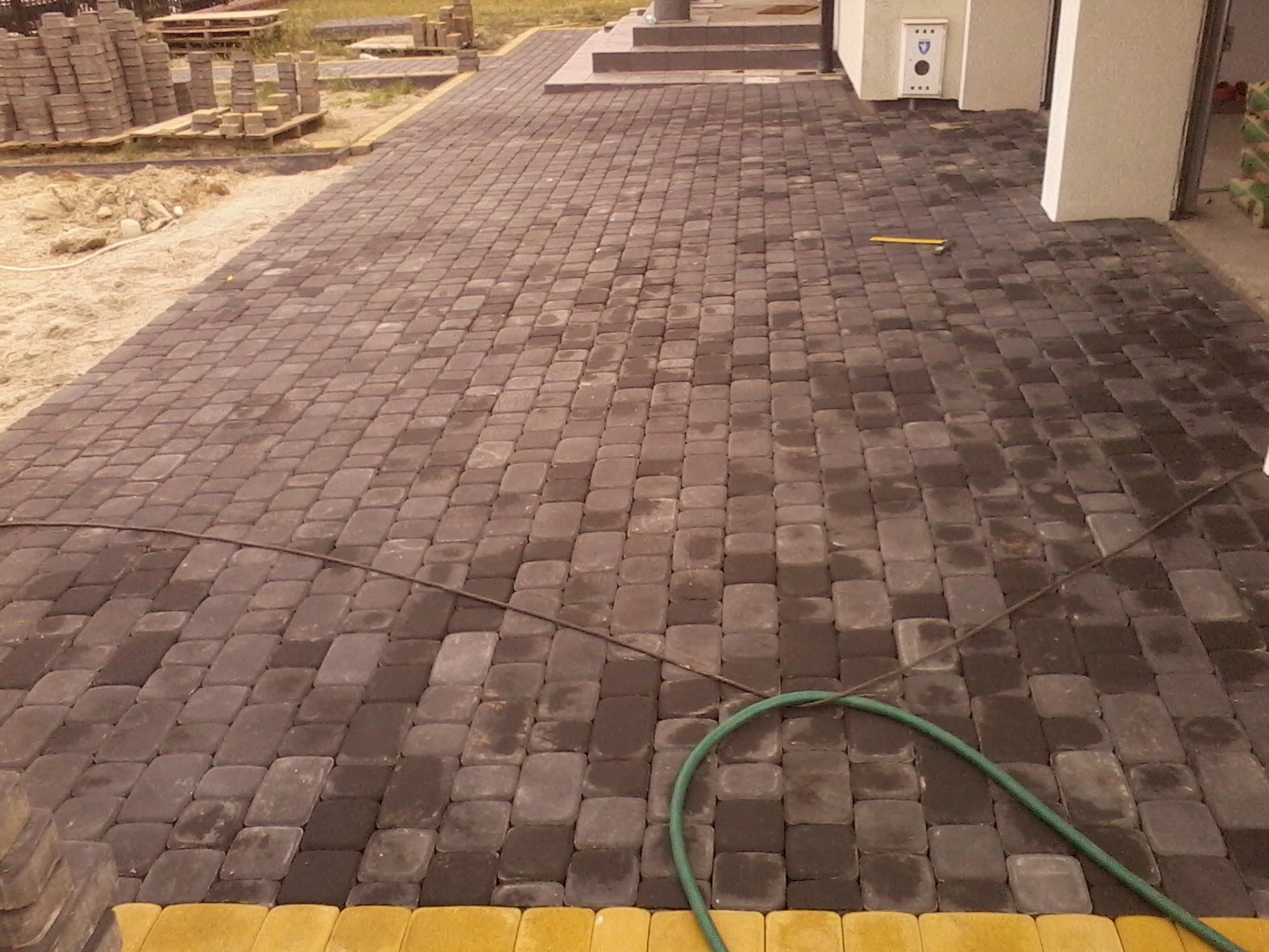 Ułożona kostka brukowa w Bydgoszczy, widoczny fragment węża ogrodowego i narzędzie, w tle palety z kostką oraz fragment budynku.