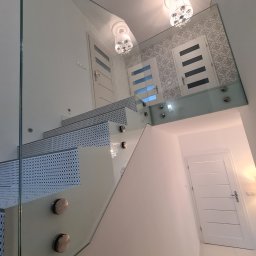 Wojciech Gawron BALUSTRADY NIERDZEWNE - Szklana balustrada schodowa z metalowymi mocowaniami, widok z dołu, w tle drzwi i ozdobna tapeta.
