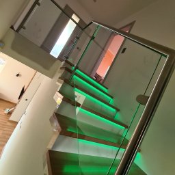 Wojciech Gawron BALUSTRADY NIERDZEWNE - Nowoczesne schody drewniane z podświetleniem LED na stopniach i szklaną balustradą z nierdzewnymi elementami mocującymi, widok z dołu pod kątem.