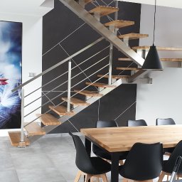 Wojciech Gawron BALUSTRADY NIERDZEWNE - Nowoczesne schody z drewnianymi stopniami i metalową balustradą w jasnym wnętrzu z szarą ścianą akcentującą, obok stół z czarnymi krzesłami.