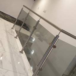 Wojciech Gawron BALUSTRADY NIERDZEWNE - Nowoczesne schody z balustradą ze stali nierdzewnej i szklanymi panelami, widoczne białe marmurowe płytki podłogowe i elementy instalacji elektrycznej na ścianie.