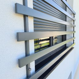 Wojciech Gawron BALUSTRADY NIERDZEWNE - Szare, metalowe zabezpieczenie okna z poziomych i pionowych profili, zamontowane na elewacji budynku z roletą zewnętrzną.