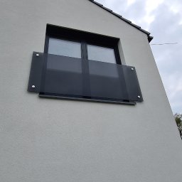 Wojciech Gawron BALUSTRADY NIERDZEWNE - Szklana balustrada balkonowa zamontowana na elewacji budynku z widocznym oknem i fragmentem dachu.