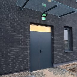 Wojciech Gawron BALUSTRADY NIERDZEWNE - Nowoczesne, grafitowe drzwi wejściowe do budynku z ciemnej cegły, osłonięte szklanym daszkiem na metalowej konstrukcji, z oświetleniem awaryjnym nad wejściem.