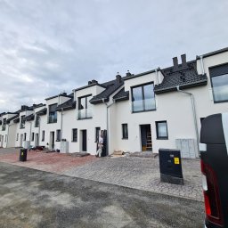 Wojciech Gawron BALUSTRADY NIERDZEWNE - Szereg nowo wybudowanych, białych domów szeregowych z czarnymi dachami i oknami, z pracownikiem montującym szklaną balustradę balkonową. Widoczny fragment chodnika z kostki brukowej i samochód...