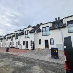 Szereg nowo wybudowanych, białych domów szeregowych z czarnymi dachami i oknami, z pracownikiem montującym szklaną balustradę balkonową. Widoczny fragment chodnika z kostki brukowej i samochód...