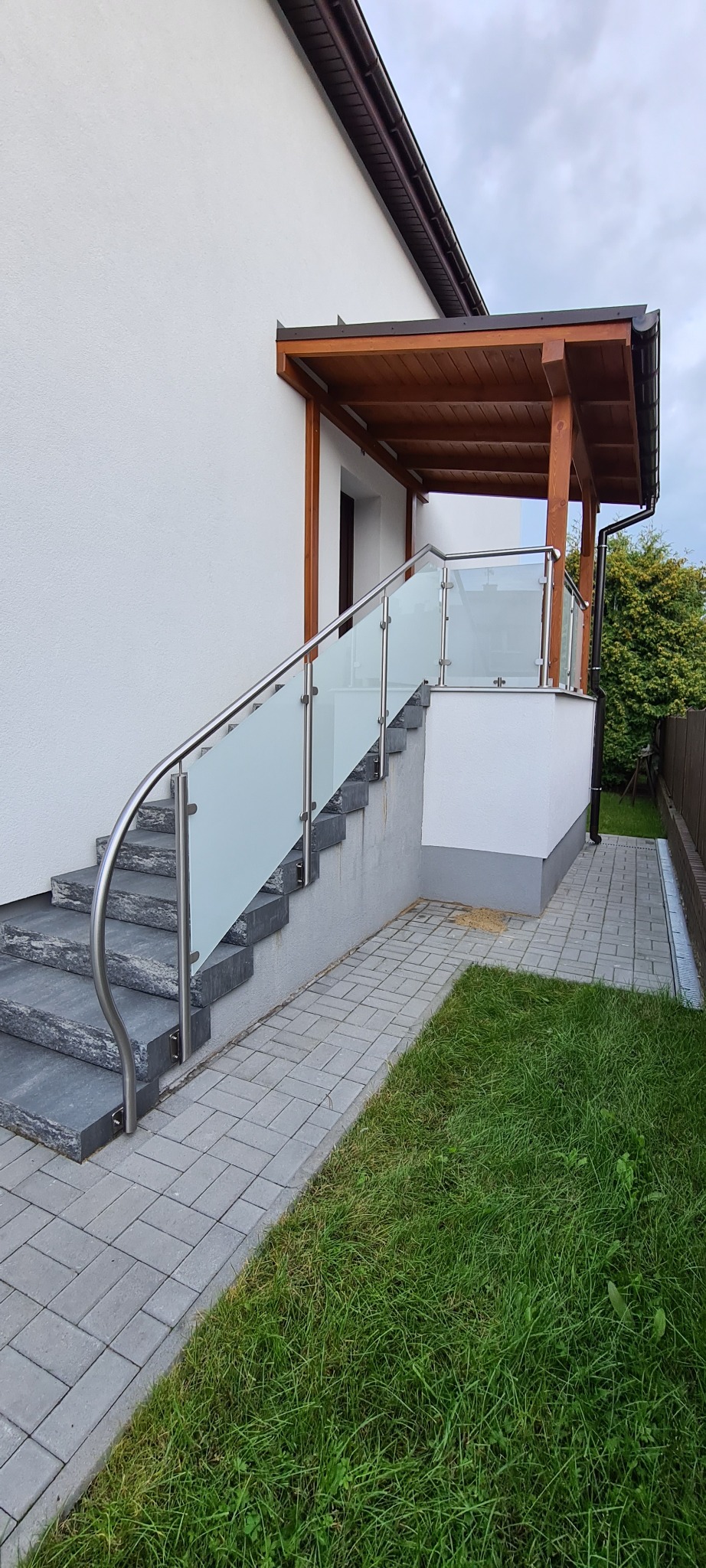 Zewnętrzne schody z szarego kamienia prowadzące do zadaszonego wejścia, zabezpieczone nowoczesną balustradą ze stali nierdzewnej z wypełnieniem z matowego szkła.