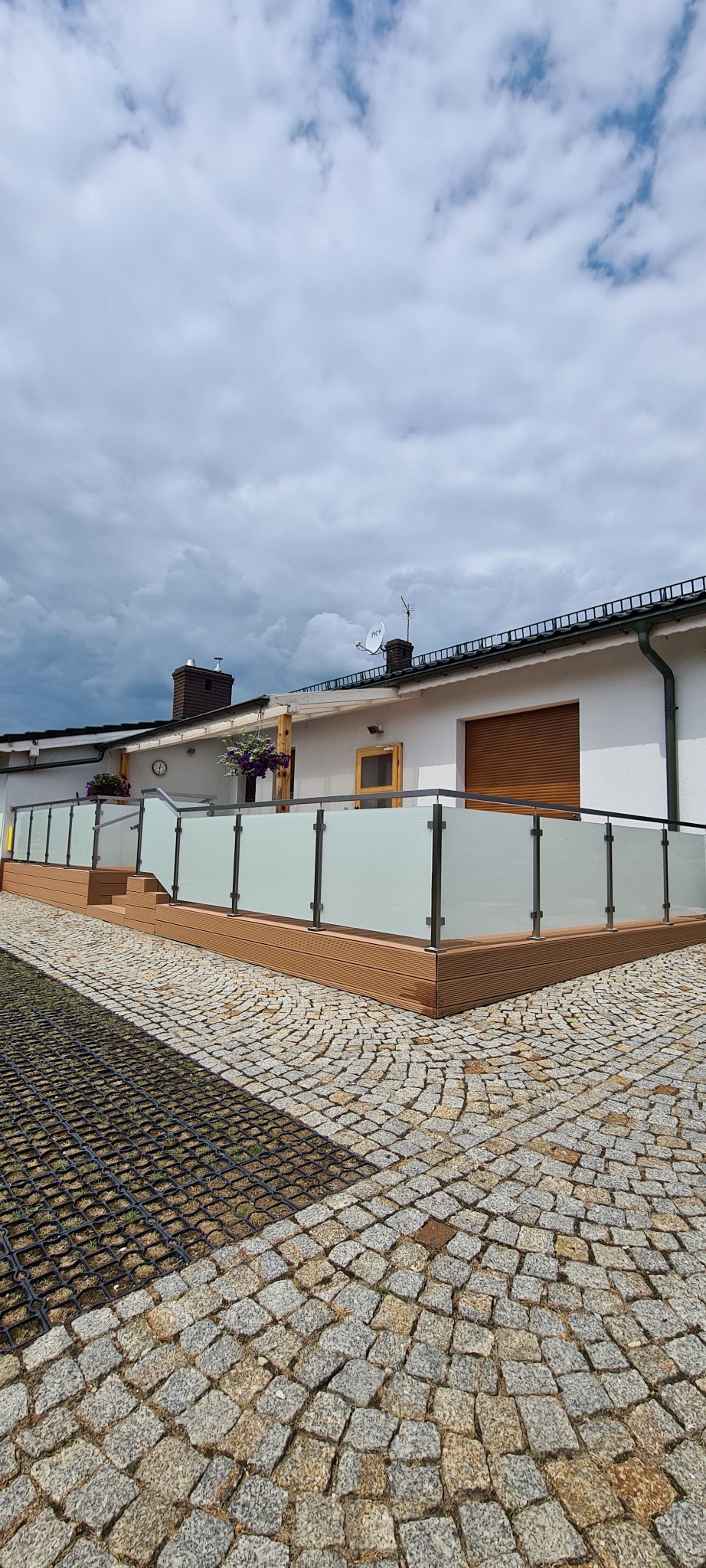 Nowoczesna balustrada ze szkła matowego i stali nierdzewnej na tarasie z drewnianą podłogą, widok z poziomu brukowanej ścieżki.