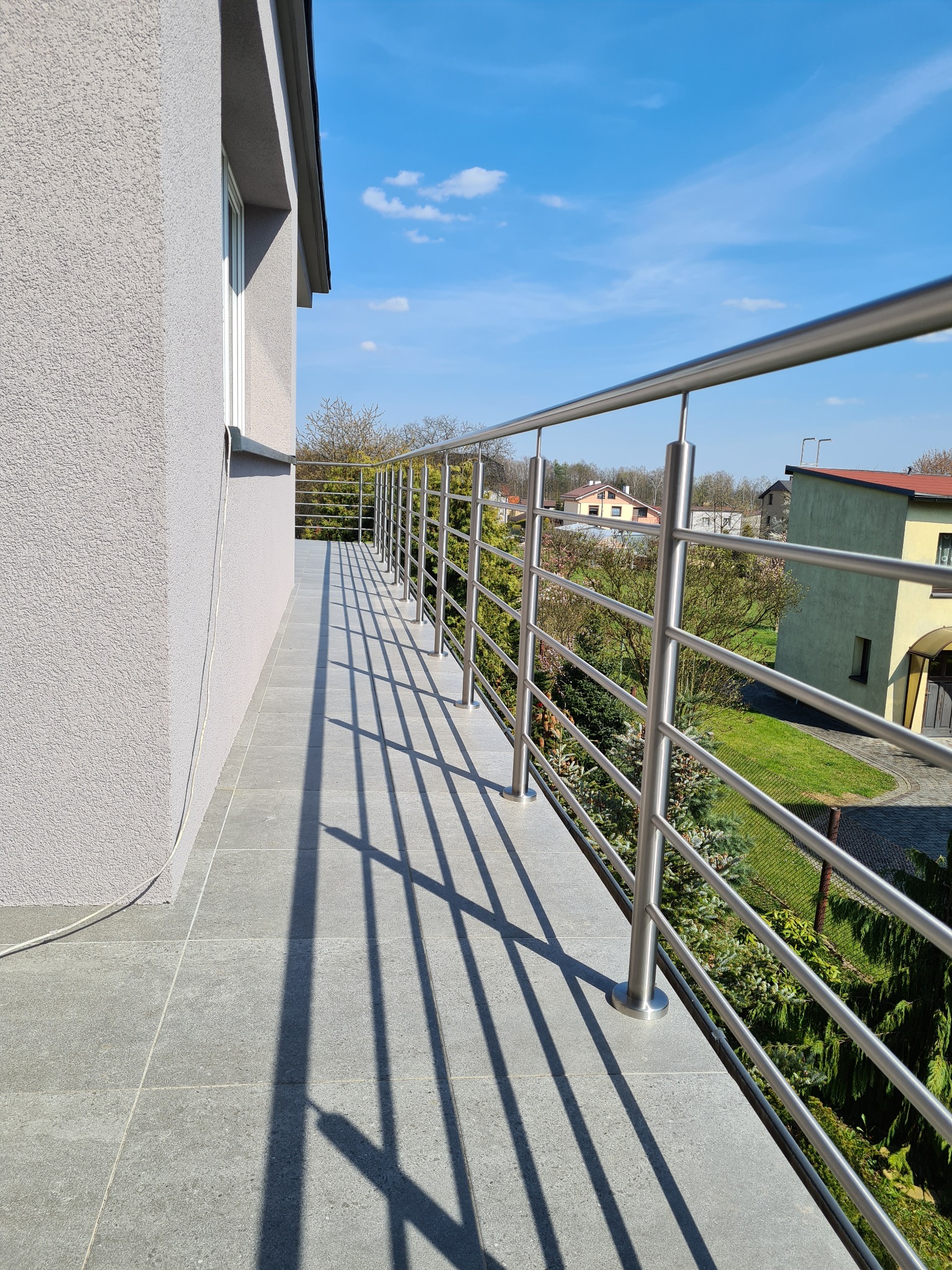 Balkon z nowoczesną, metalową balustradą z rurek, rzucającą wyraźne cienie na szare płytki podłogowe. W tle widoczne budynki i zieleń w słoneczny dzień.