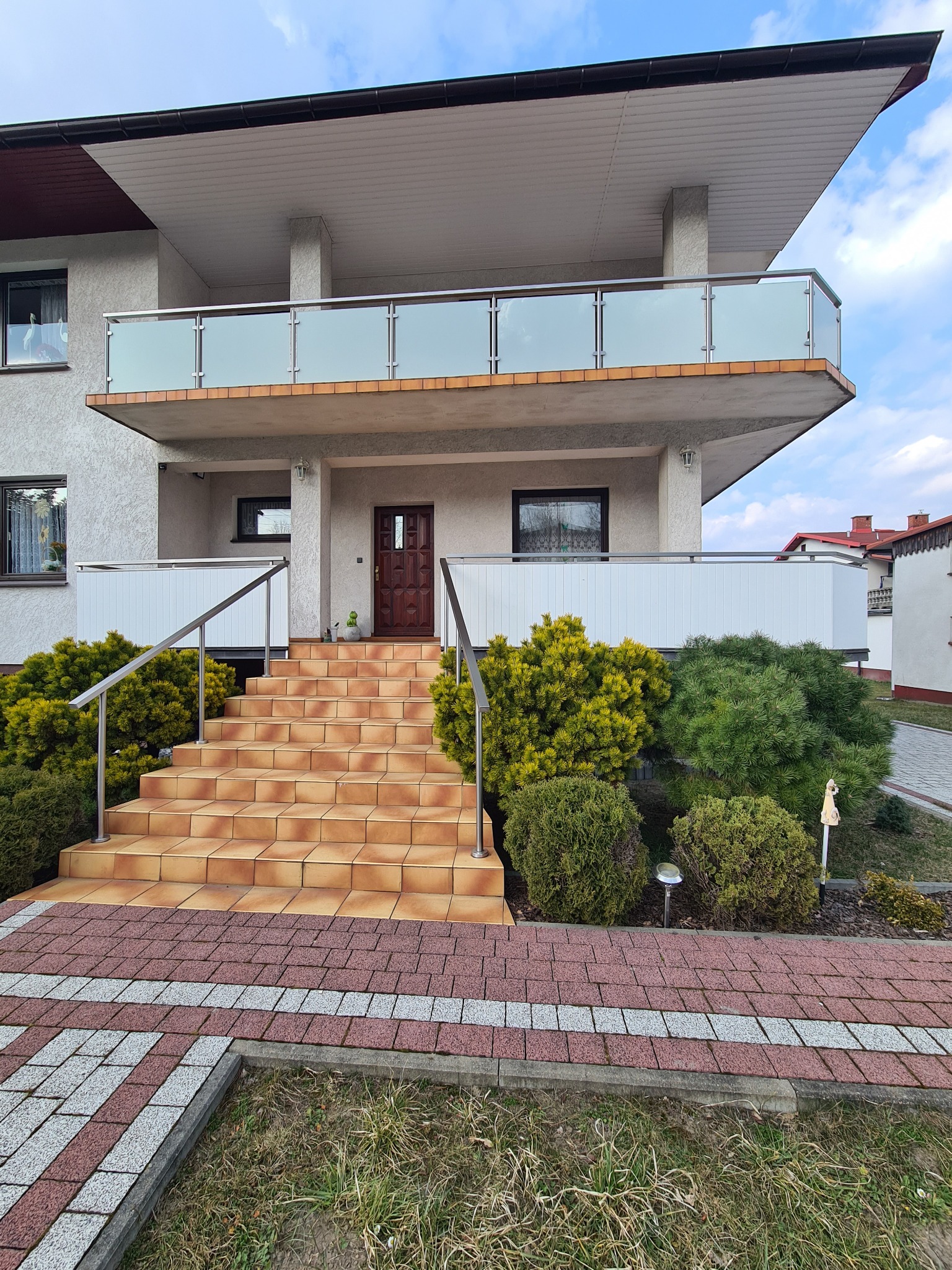 Nowoczesny dom w Koszęcinie z balkonem zabezpieczonym balustradą ze szkła i stali nierdzewnej oraz schodami wejściowymi z poręczami.