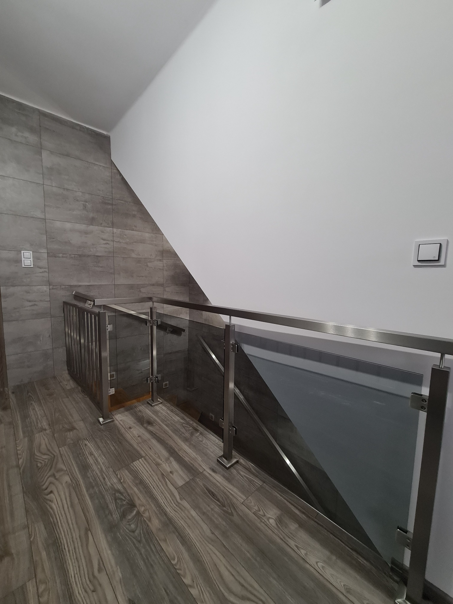 Szklana balustrada ze stali nierdzewnej z poziomymi elementami, zamontowana przy schodach w nowoczesnym wnętrzu z szarymi płytkami na ścianie i podłodze imitującej drewno.