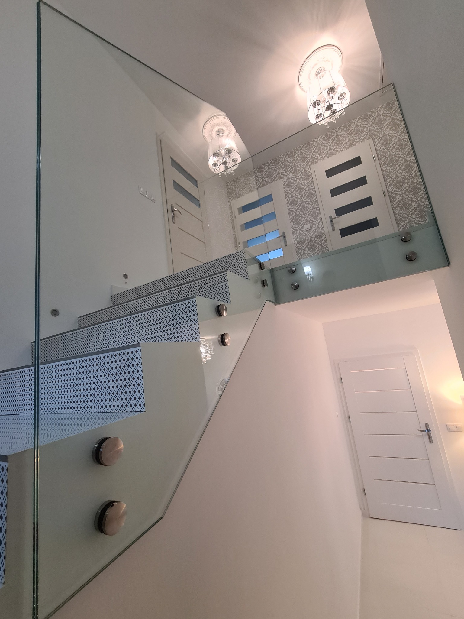 Szklana balustrada schodowa z metalowymi mocowaniami, widok z dołu, w tle drzwi i ozdobna tapeta.