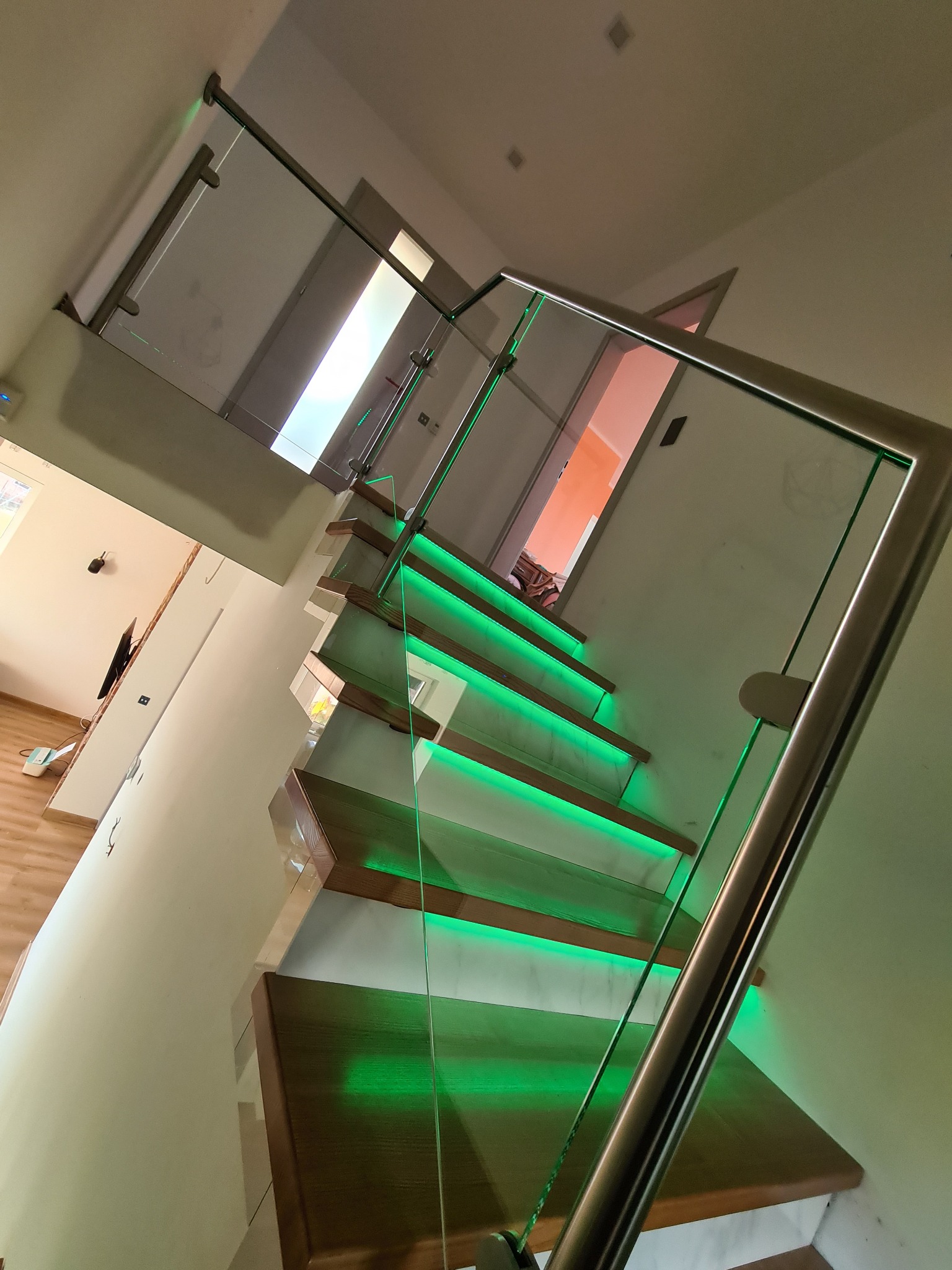 Nowoczesne schody drewniane z podświetleniem LED na stopniach i szklaną balustradą z nierdzewnymi elementami mocującymi, widok z dołu pod kątem.