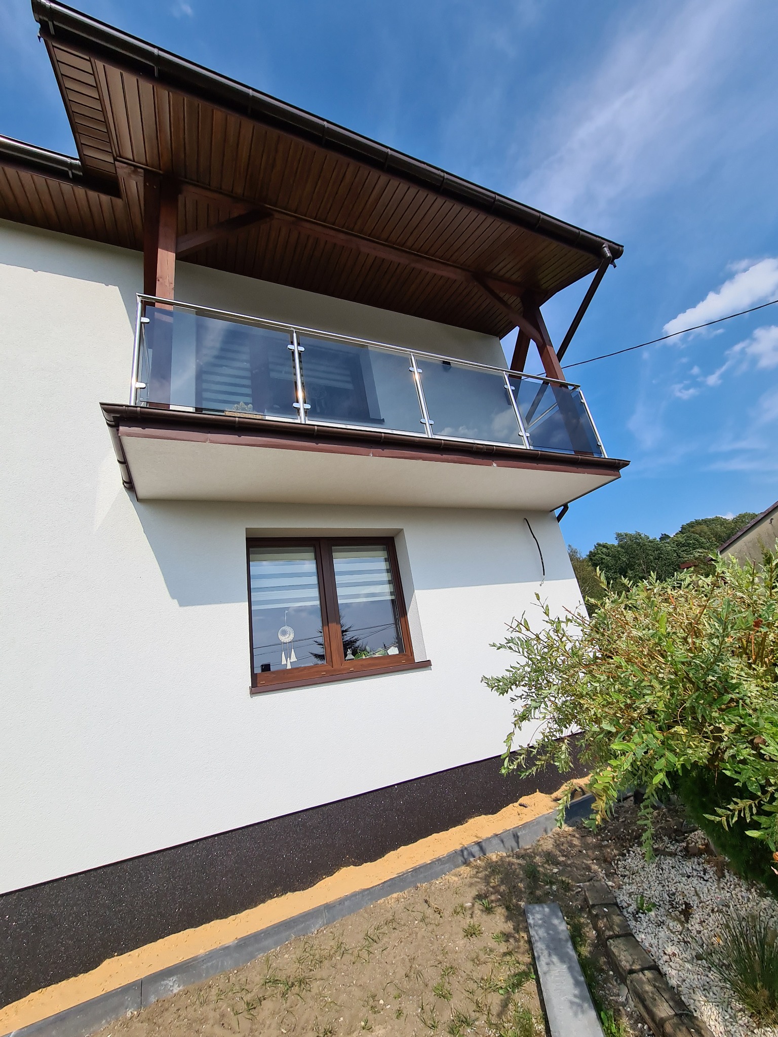 Nowoczesny balkon z balustradą nierdzewną i szklanymi panelami, drewniana konstrukcja zadaszenia, biała elewacja budynku w słoneczny dzień.