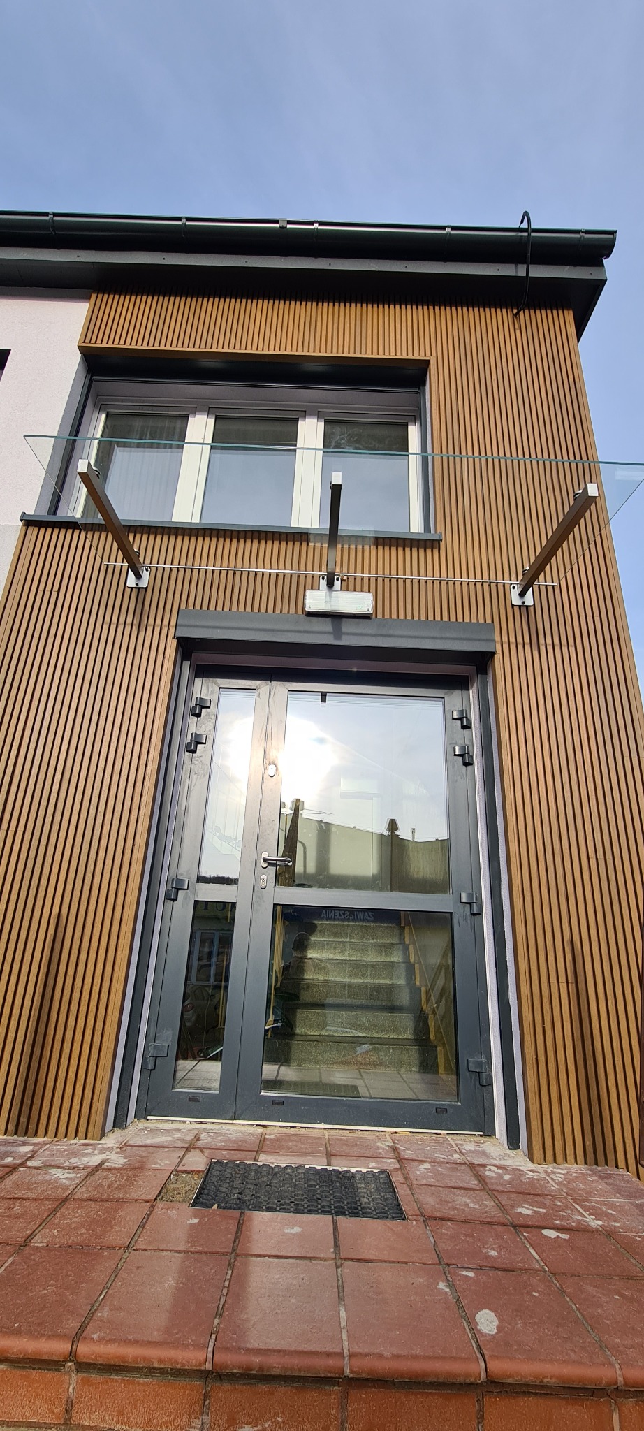 Nowoczesne wejście do domu z drewnianą elewacją, szklanym zadaszeniem wspartym na metalowych wspornikach i szarymi drzwiami wejściowymi z widocznymi schodami wewnątrz.