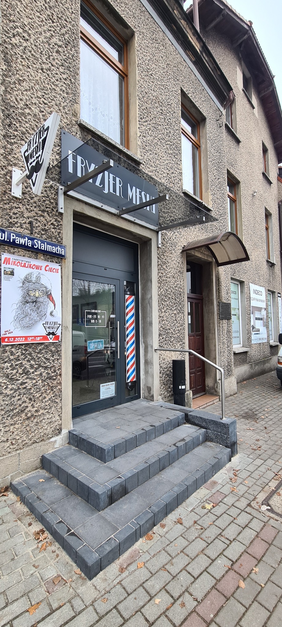 Wejście do salonu fryzjerskiego z trzema stopniami z szarej kostki, balustradą ze stali nierdzewnej i charakterystycznym barber pole obok szklanych drzwi wejściowych.
