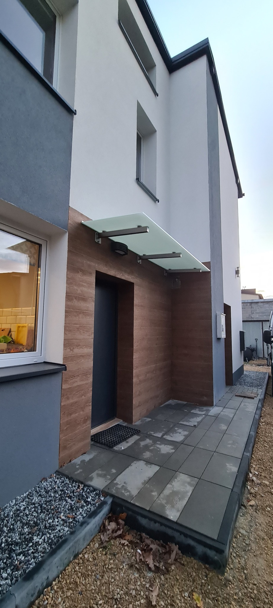 Nowoczesne wejście do domu z szarą elewacją, drewnopodobną okładziną i szklanym zadaszeniem wspartym na metalowych wspornikach, wyłożone szarą kostką brukową.