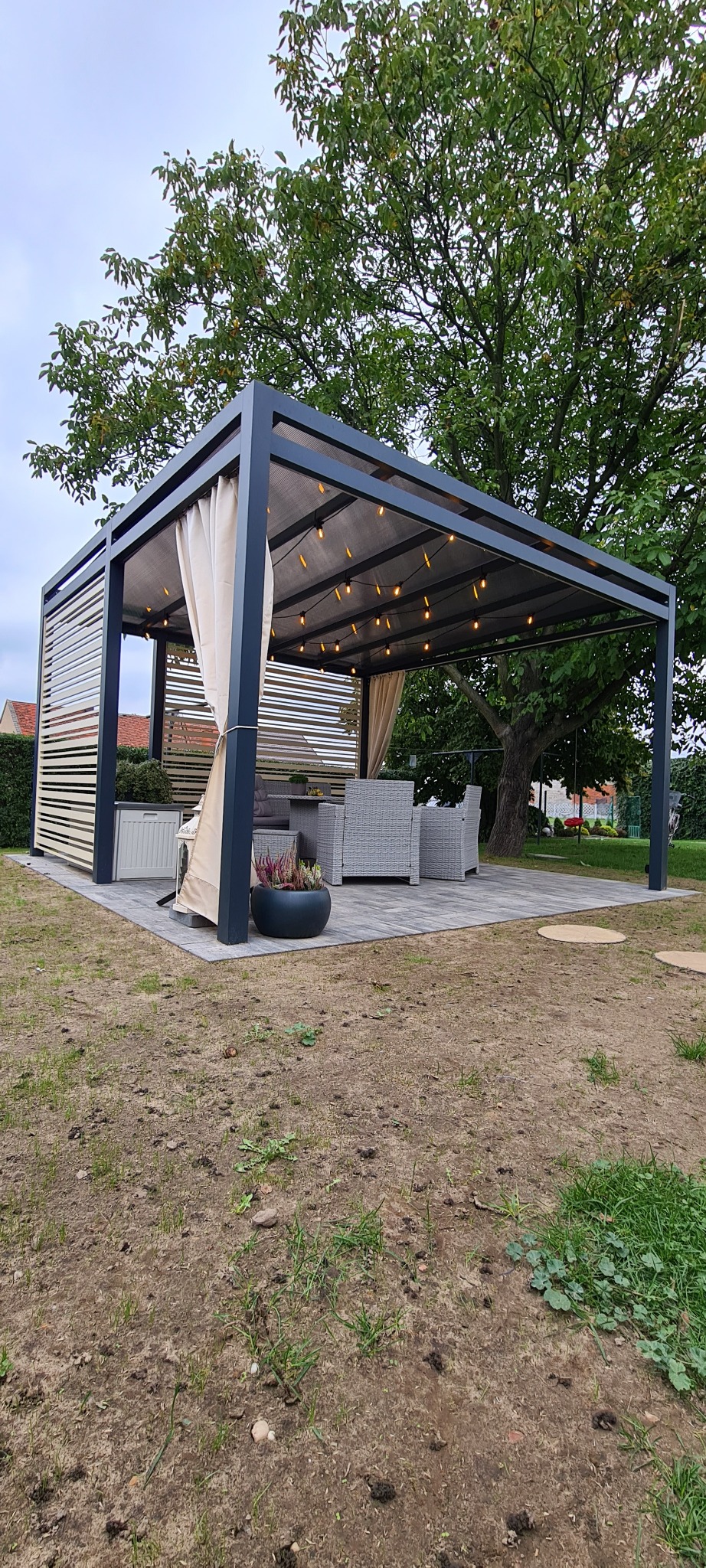 Nowoczesna pergola z szarymi metalowymi słupkami, drewnianymi panelami, jasnymi zasłonami i oświetleniem w postaci girlandy żarówek, ustawiona na wybrukowanym patio w ogrodzie.