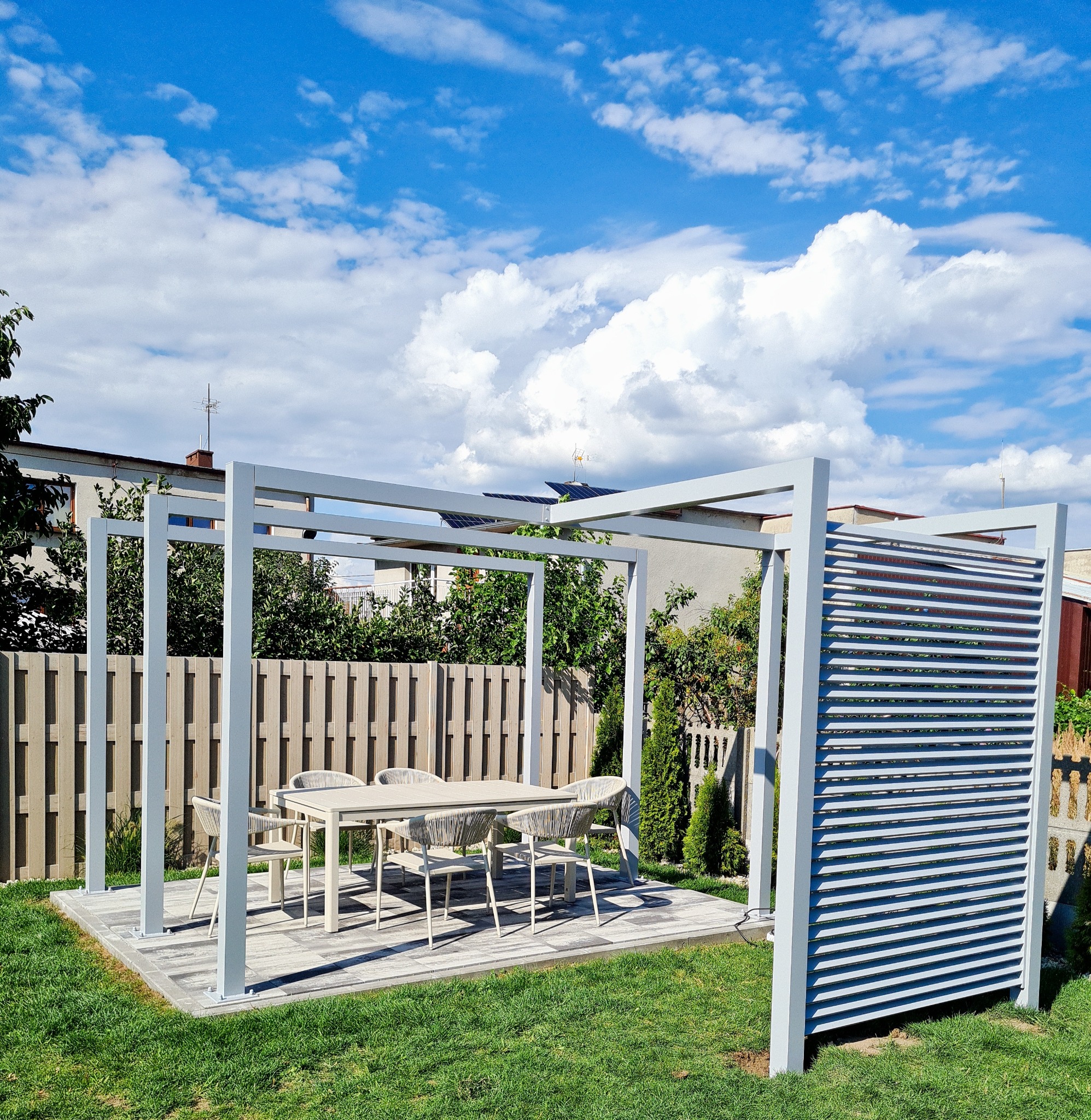 Nowoczesna pergola z białymi, metalowymi słupami i lamelami, wyposażona w stół i krzesła z plecionki, na tle trawnika i niebieskiego nieba z chmurami.