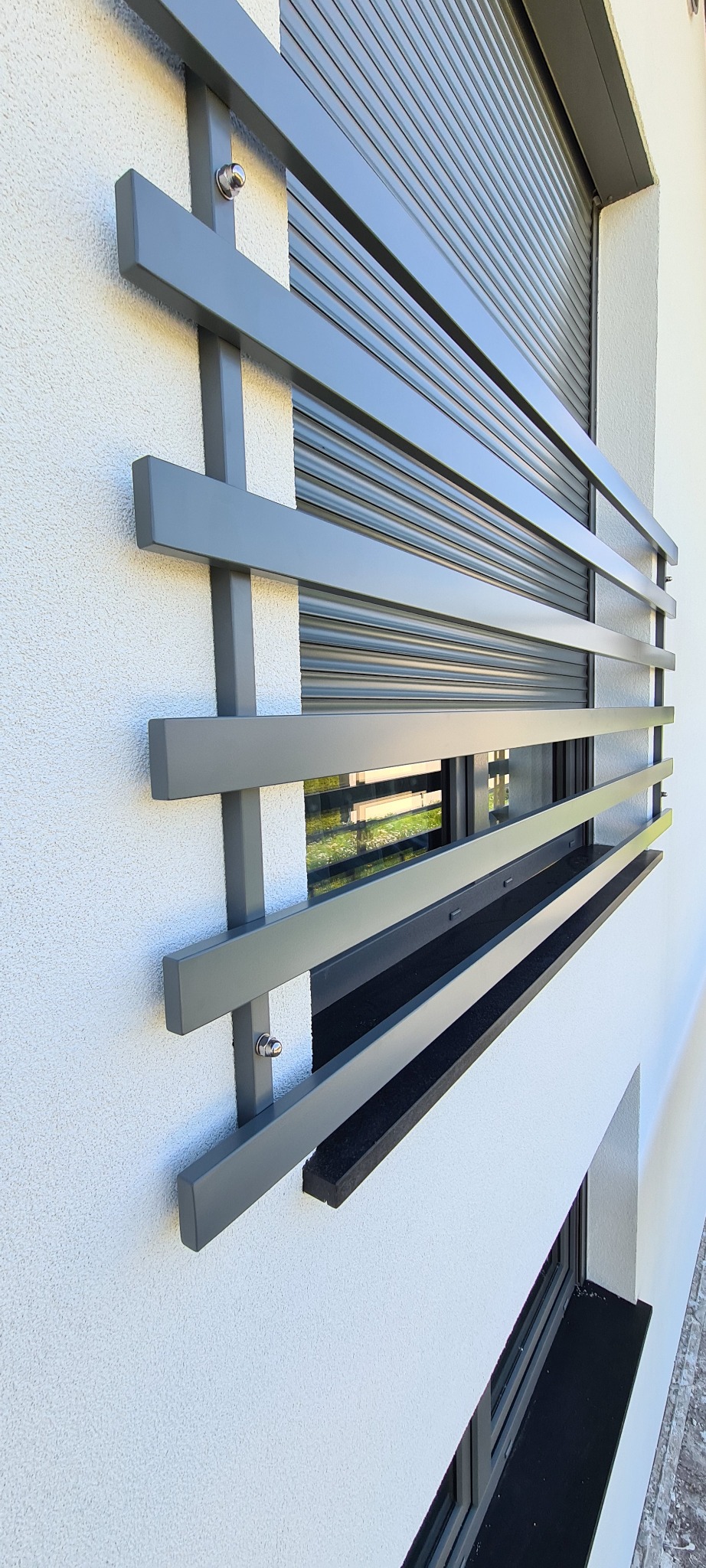 Szare, metalowe zabezpieczenie okna z poziomych i pionowych profili, zamontowane na elewacji budynku z roletą zewnętrzną.