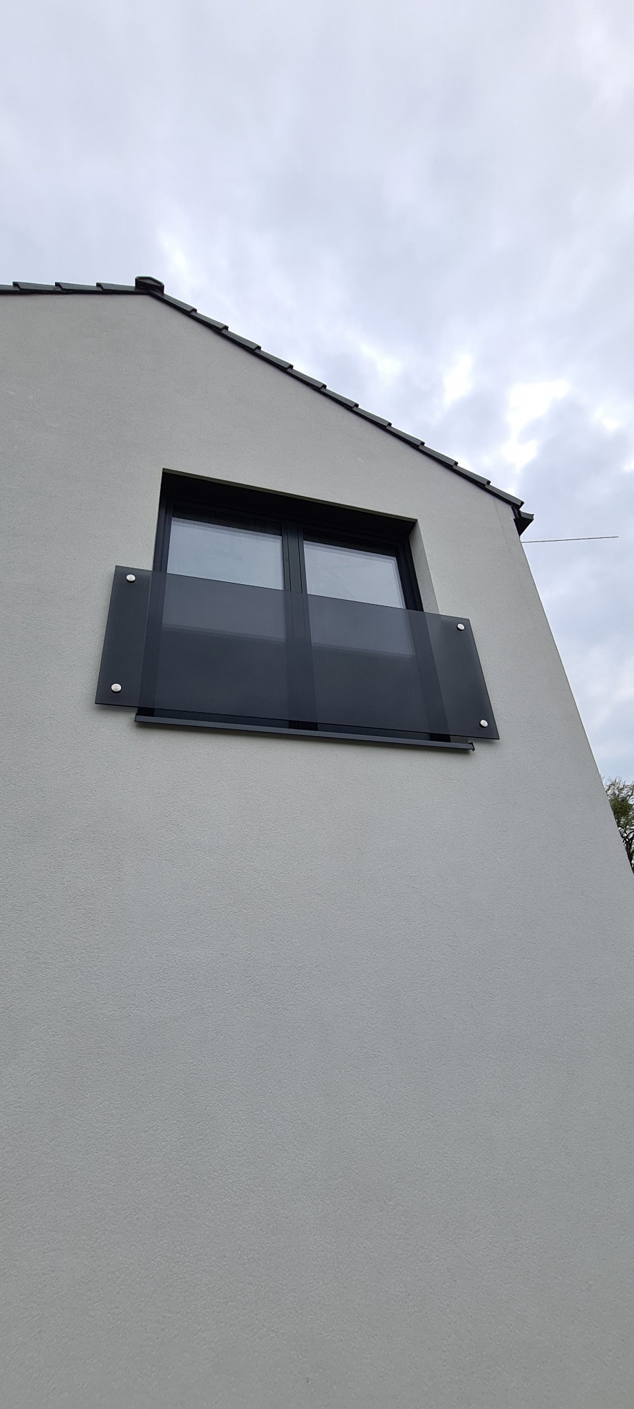 Szklana balustrada balkonowa zamontowana na elewacji budynku z widocznym oknem i fragmentem dachu.