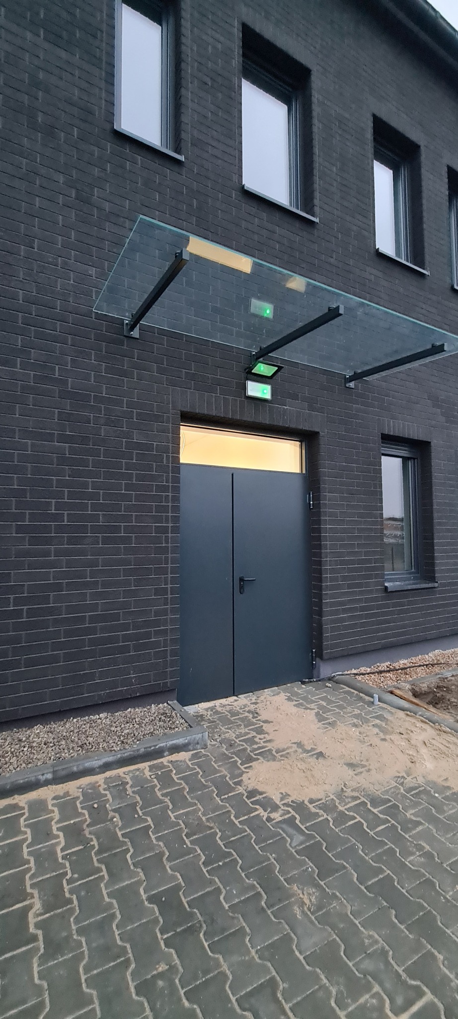 Nowoczesne, grafitowe drzwi wejściowe do budynku z ciemnej cegły, osłonięte szklanym daszkiem na metalowej konstrukcji, z oświetleniem awaryjnym nad wejściem.