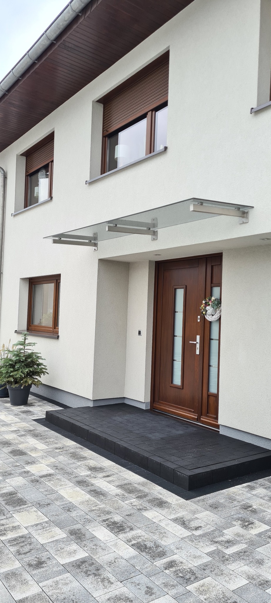 Zadaszenie ze szkła hartowanego nad brązowymi drzwiami wejściowymi z dekoracyjnym wieńcem, widok z zewnątrz na tle jasnej elewacji domu z oknami i roletami.