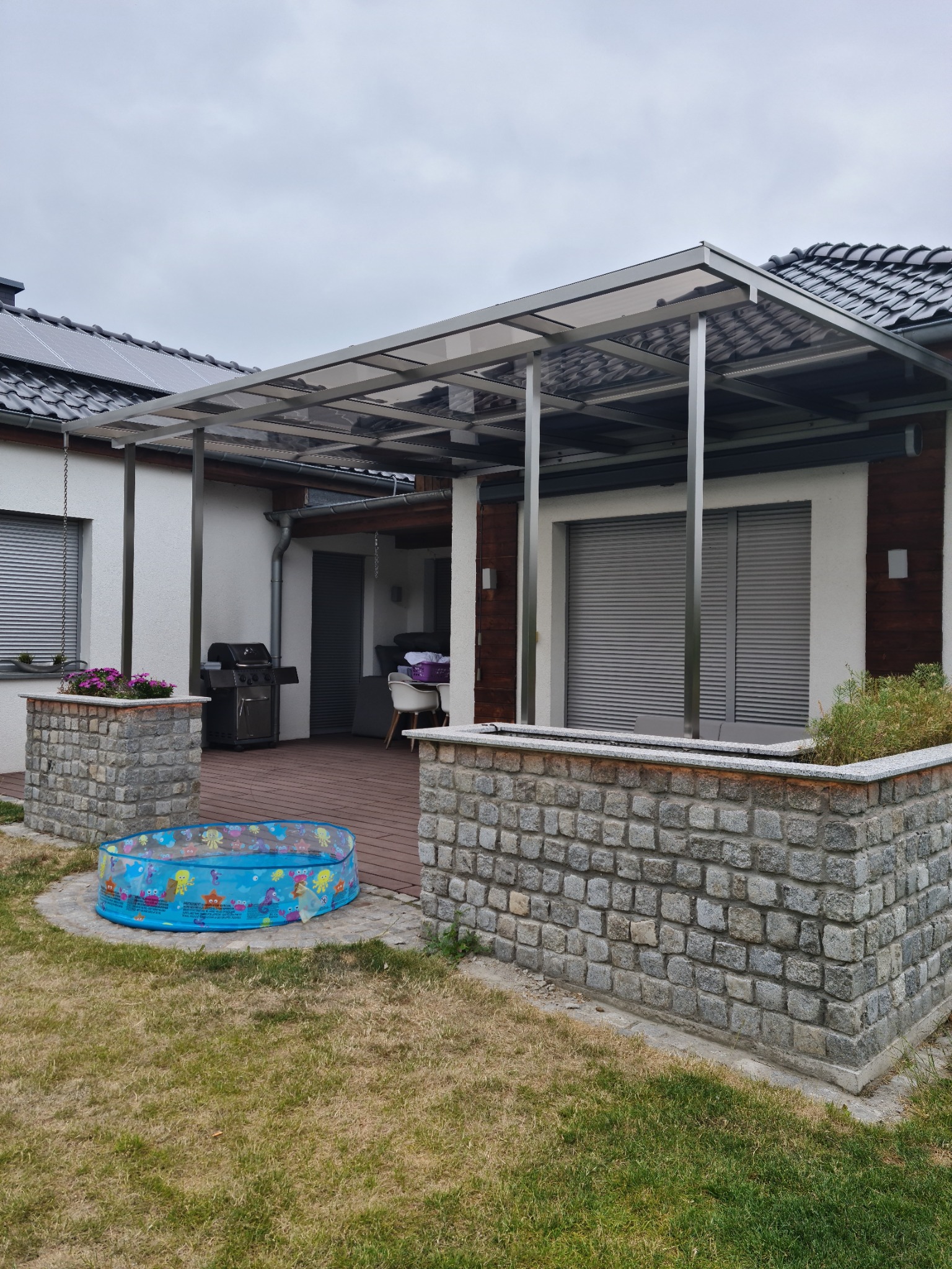 Zadaszenie tarasu z poliwęglanu na metalowej konstrukcji, widok z ogrodu, na pierwszym planie mały basenik dziecięcy.