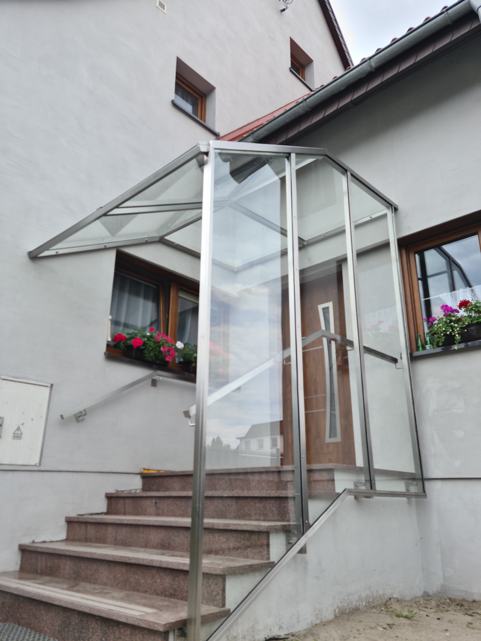Zadaszone wejście do domu z przeszkloną konstrukcją wspartą na metalowych elementach, prowadzące po schodach z brązowego kamienia, z widocznymi donicami z kwiatami w oknach.