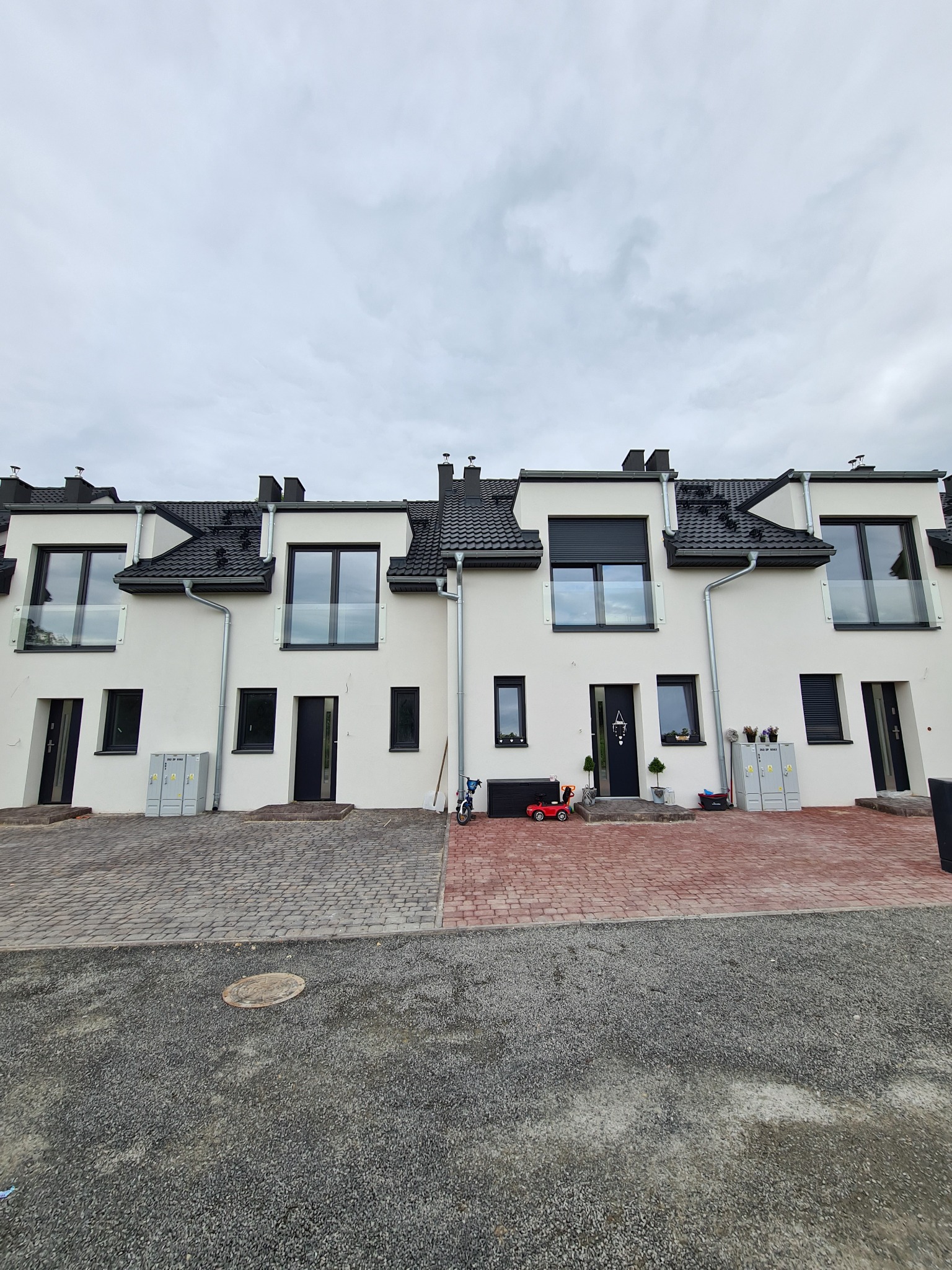 Nowe, białe domy szeregowe z czarnymi dachami i oknami, balustrady balkonowe ze szkła, kostka brukowa przed wejściami, pochmurne niebo.