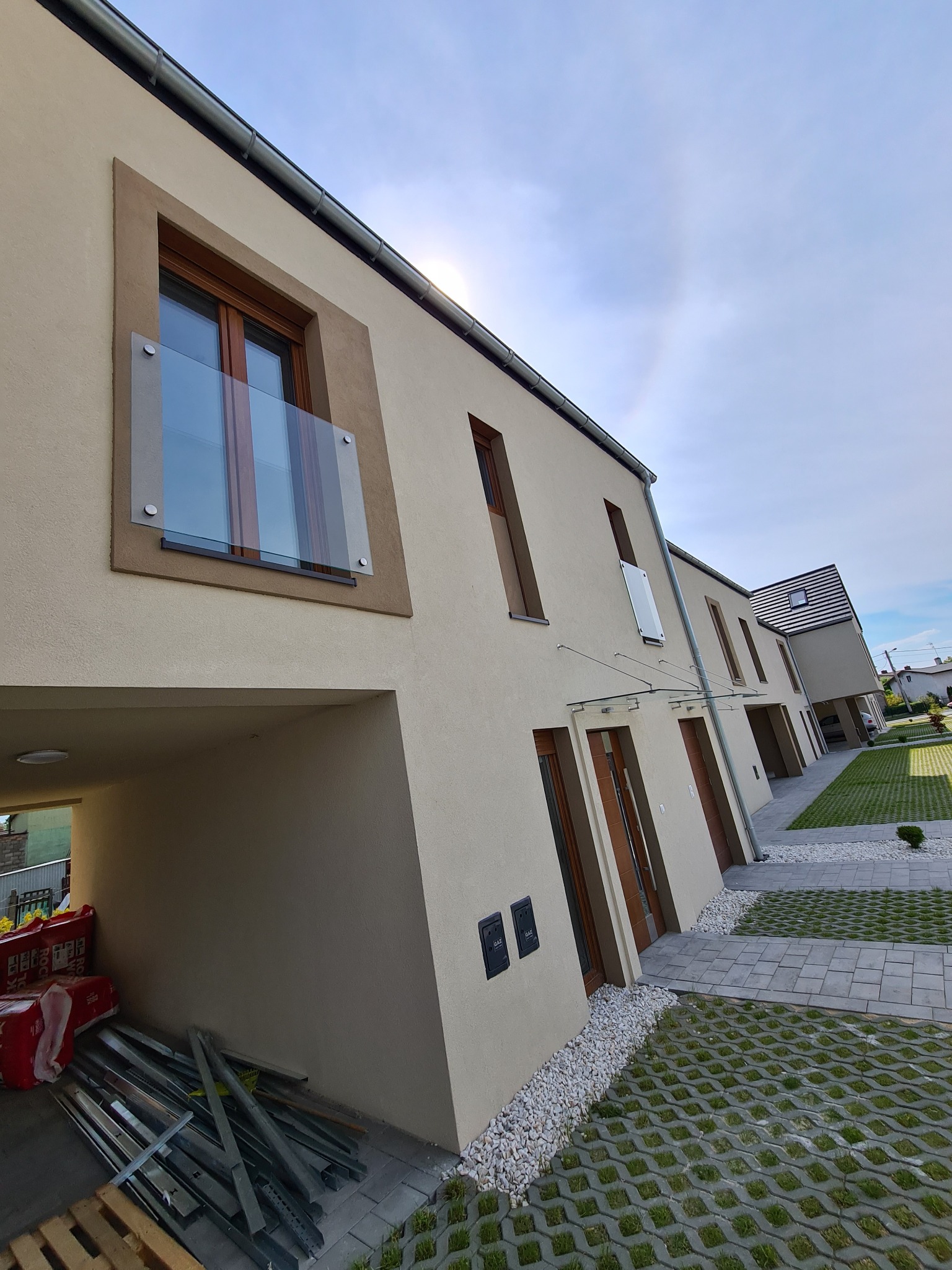 Nowoczesne szeregówki z szklanymi balustradami balkonowymi i zadaszeniem nad wejściem, widok z boku na uporządkowane otoczenie z ażurowymi płytami trawnikowymi i stertą materiałów budowlanych...